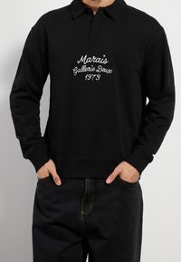 Sort langærmet polo med krave, med hvid broderet tekst "Marais Gallerie Deux 1973." Stoffet virker blødt med ribstrikkede manchetter.