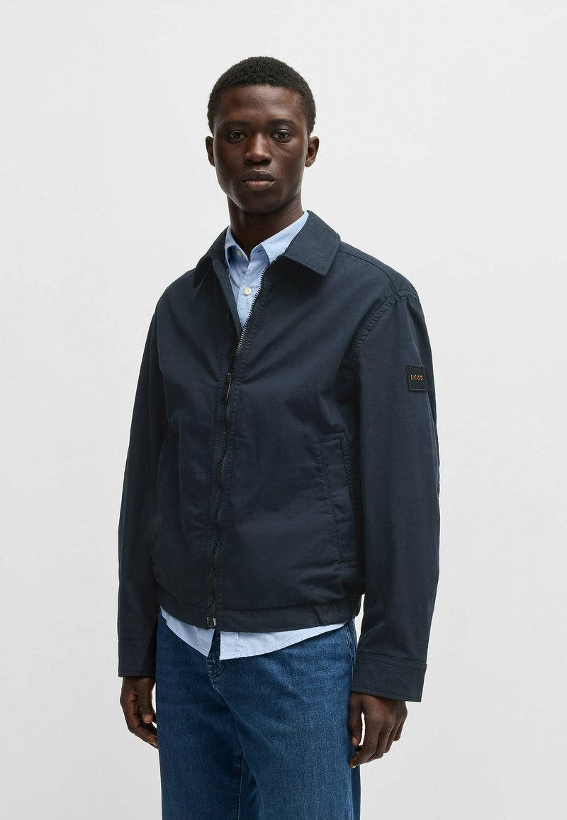 Giacca leggera di colore blu navy con zip frontale, colletto e tasche laterali. Materiale in misto cotone con una texture morbida.