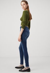 Mørkeblå, skinny jeans med frynset kant, kombinert med en tettsittende olivengrønn genser med lange ermer. Svarte sko med strukturert mønster og sokker med prikker.