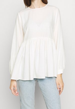 Kobieta w luźnej, białej bluzce z długimi rękawami, z marszczeniem w talii, w jasnoniebieskich jeansach na tle jednolitego tła.