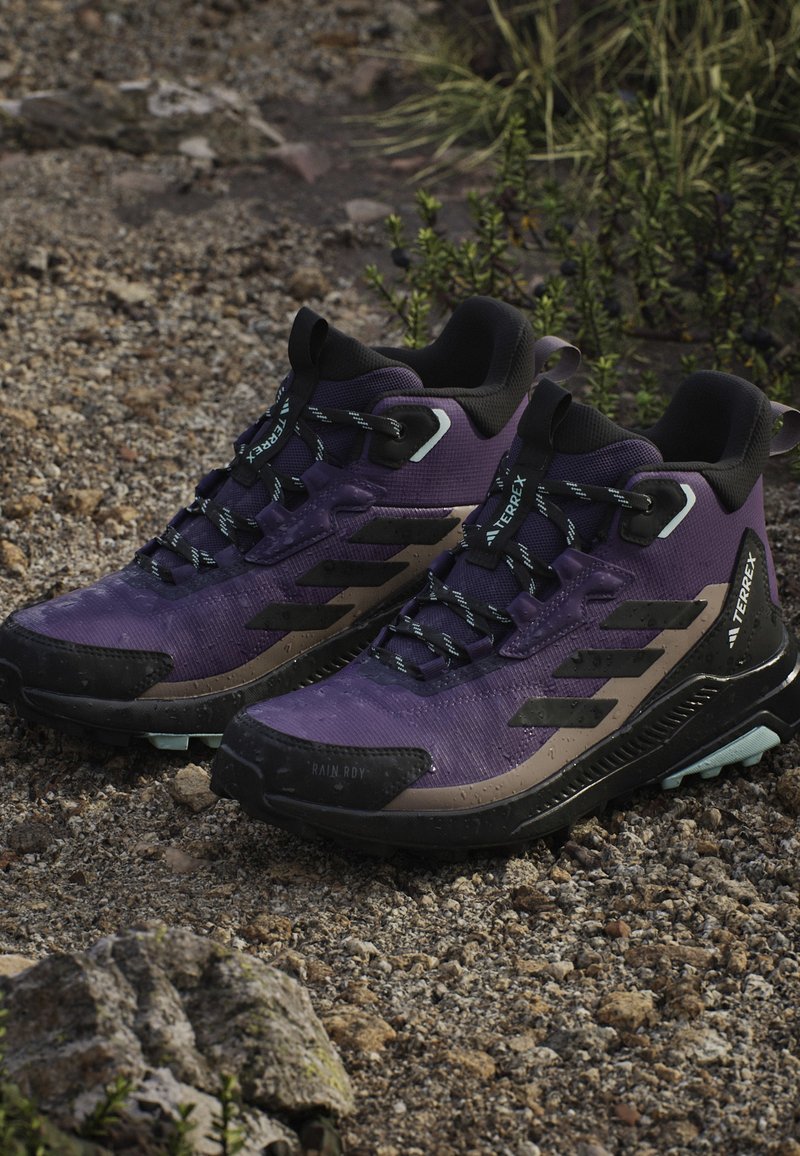 Botas de senderismo moradas con acentos en gris oscuro, parte superior de sintético texturizado, suela de goma y cordones reflectantes. Características impermeables destacadas.