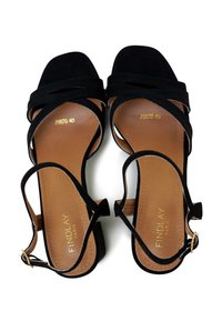 Black suede heeled sandals with crisscross straps, open toe, and a tan insole featuring gold embossed branding. Adjustable ankle strap detail.