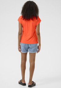 Short-sleeve orange top paired with denim shorts featuring rolled cuffs. Model stands barefoot, showcasing multi-textured fabric and relaxed fit.