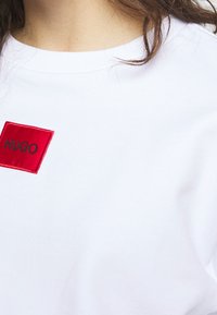 Camiseta de algodón blanca con una etiqueta de tela roja con "HUGO" bordado en negro. Diseño sencillo de cuello redondo. Textura suave, estilo casual.