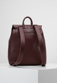 Zaino in pelle marrone scuro con forma strutturata, dotato di spallacci regolabili e manico superiore, texture liscia e hardware minimo.