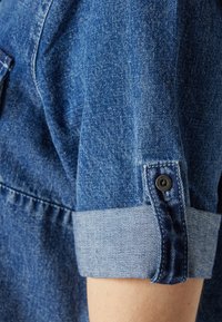 Jeansskjorta med en ljusblå manschett, uppvikta ärmar och en slät textur. Har en liten knappdetalj vid manschetten för säker stängning.