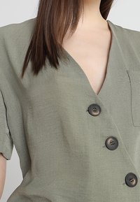 Olivgrön linbluse med korta ärmar, V-ringning och tre svarta knappar fram. Har en bröstficka på vänster sida.