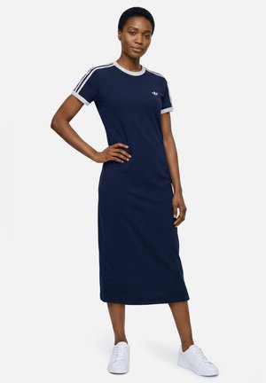 Vrouw draagt een donkerblauwe Adidas midi-jurk met witte strepen op de mouwen en witte sneakers, staand met één hand op haar heup.