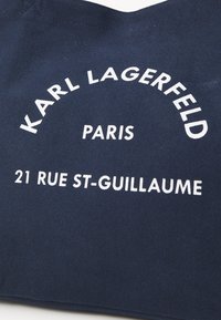 Marineblauwe canvas tas met witte tekst: "KARL LAGERFELD" gebogen boven "PARIJS" en "21 RUE ST-GUILLAUME" onderaan. De tekst is vetgedrukt en duidelijk.