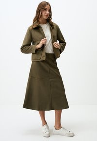 Olivgrüne Jacke mit strukturiertem Kragen, kombiniert mit einem passenden Rock in fließender Silhouette und Nahtdetails. Weiße Sneakers sind inklusive.