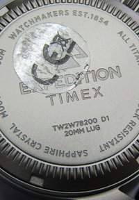 Fond de montre en titane argenté avec texte gravé : "Expédition Timex," numéro de modèle, et "Cristal Saphir." Forme circulaire avec bords texturés.