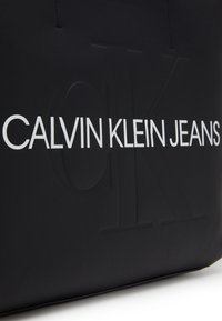 Svart toteväska med präglad "CALVIN KLEIN JEANS"-logotyp i vitt, tillverkad av slätt material, med hållbara handtag och rektangulär form.