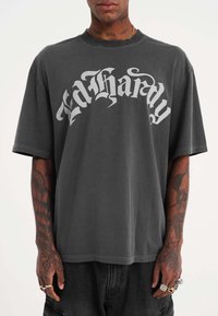 Camiseta gris de gran tamaño hecha de algodón, con un gran logo en blanco que dice "Ed Hardy" en el pecho. Tiene mangas cortas y cuello redondo.