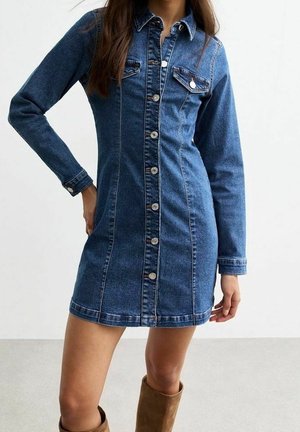 Kvinde iført en tætsiddende blå denim knap-på kjole med lange ærmer og brystlommer, kombineret med brune knæhøje støvler.