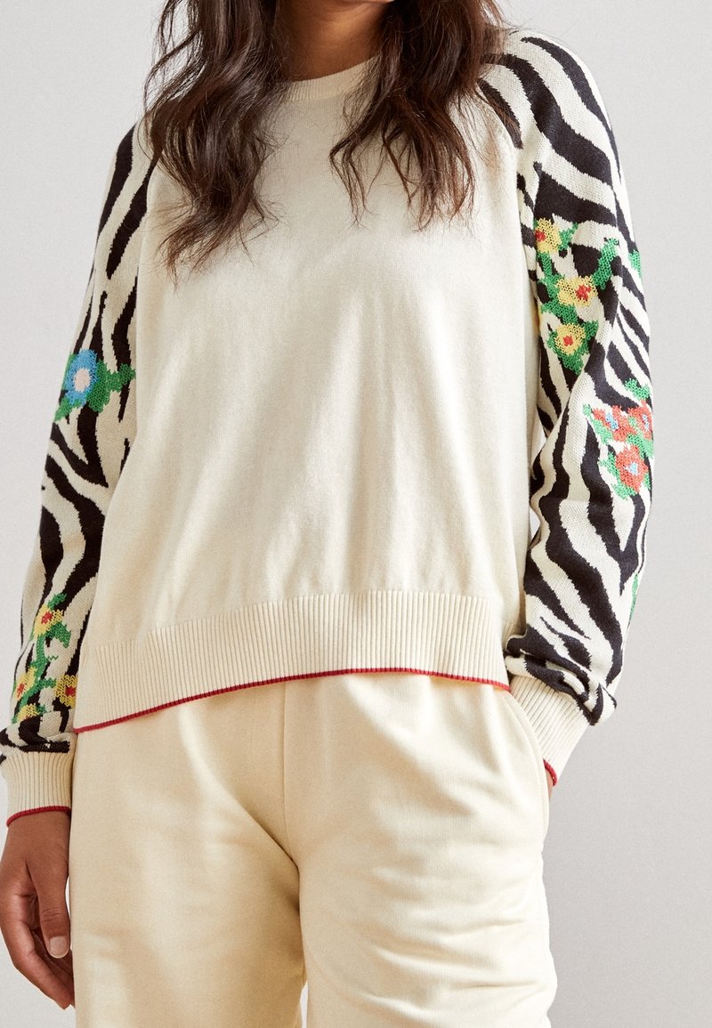 Femme portant un pull crème avec des manches imprimées zèbre noir et blanc décorées de fleurs brodées colorées, associé à un pantalon crème.