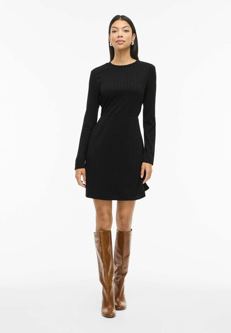 Robe noire à manches longues côtelées avec une taille cintrée et une jupe évasée. Portée avec des bottes marron montant jusqu'aux genoux, créant une silhouette élégante.