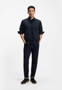 Camicia blu navy con bottoni e piccoli motivi a triangolo bianco, indossata sopra una t-shirt blu navy. Jeans in denim scuro e scarpe da ginnastica nere con suole in gomma color marrone chiaro.