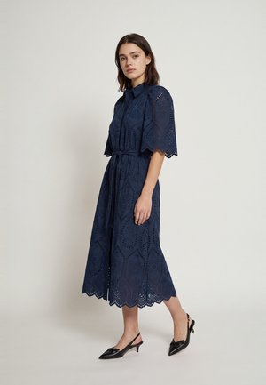 Donna che indossa un abito midi blu navy con motivo in pizzo sangallo, maniche corte, collo, cintura in vita e scarpe nere slingback con tacco kitten basso.