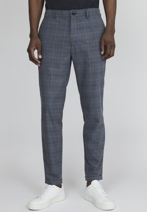 Un homme portant un pantalon slim à carreaux bleus et des baskets basses blanches, debout devant un fond gris uni.