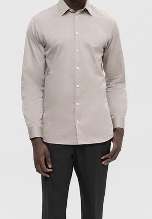 Man wearing a light beige button-up dress shirt with long sleeves and dark trousers, standing against a plain light background.