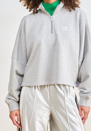 Osoba nosząca jasnoszarą bluzę Adidas z ćwiartkowym zamkiem na szyi na zielonym golfie oraz błyszczące jasnoszare spodnie z czarnymi paskami.