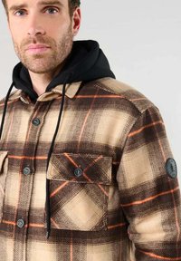 Chemise en flanelle à carreaux dans des nuances de beige, marron et orange. Elle présente deux poches à boutons, une capuche noire et un patch rond avec logo sur la manche gauche.