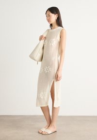 Knit beige dress with floral cutouts, side slit, and a fitted silhouette. Model holds a white bag and wears light sandals.