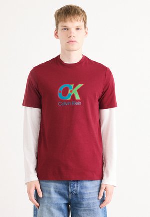 Bordeaux katoenen T-shirt met een kleurrijk "CK" logo en "Calvin Klein" tekst, overlappend een wit longsleeve shirt. Casual pasvorm.