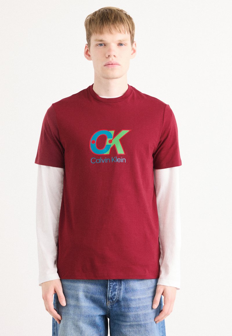 Burgunderfarget bomull T-skjorte med en fargerik "CK" logo og teksten "Calvin Klein", lagd over en hvit langermet skjorte. Casual passform.