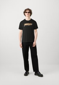 T-shirt nera in cotone con un logo grafico colorato sulla parte frontale. Abbinata a pantaloni neri e scarpe nere, il modello si trova su uno sfondo neutro.