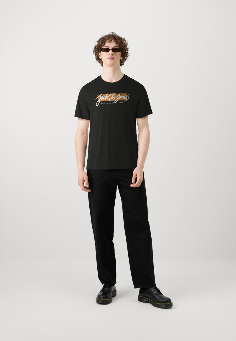 T-shirt nera in cotone con un logo grafico colorato sulla parte frontale. Abbinata a pantaloni neri e scarpe nere, il modello si trova su uno sfondo neutro.