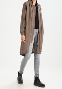 Lysbrun lang jakke med lynlås foran, ribbede manchetter og sidelommer, parret med grå slim jeans og sorte ankelstøvler.
