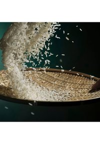 White rice grains pouring into a round woven bamboo tray held by a hand against a dark green background.
