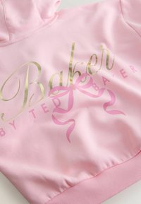 Helle rosa Kapuzenjacke mit metallic-goldenem Schriftzug "Baker" und einem pinken Schleifengrafik. Weiches Material mit geripptem Saumdetails.