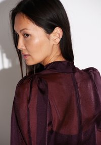 Transparante bordeaux blouse met satijnen afwerking, pofmouwen en hoog kraagdetail. Gladde textuur met subtiele glans en getailleerde snit.