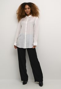 Camicia bianca a maniche lunghe con colletto, chiusura con bottoni sul davanti e maniche con polsini, abbinata a pantaloni neri larghi e scarpe nere con tacco.