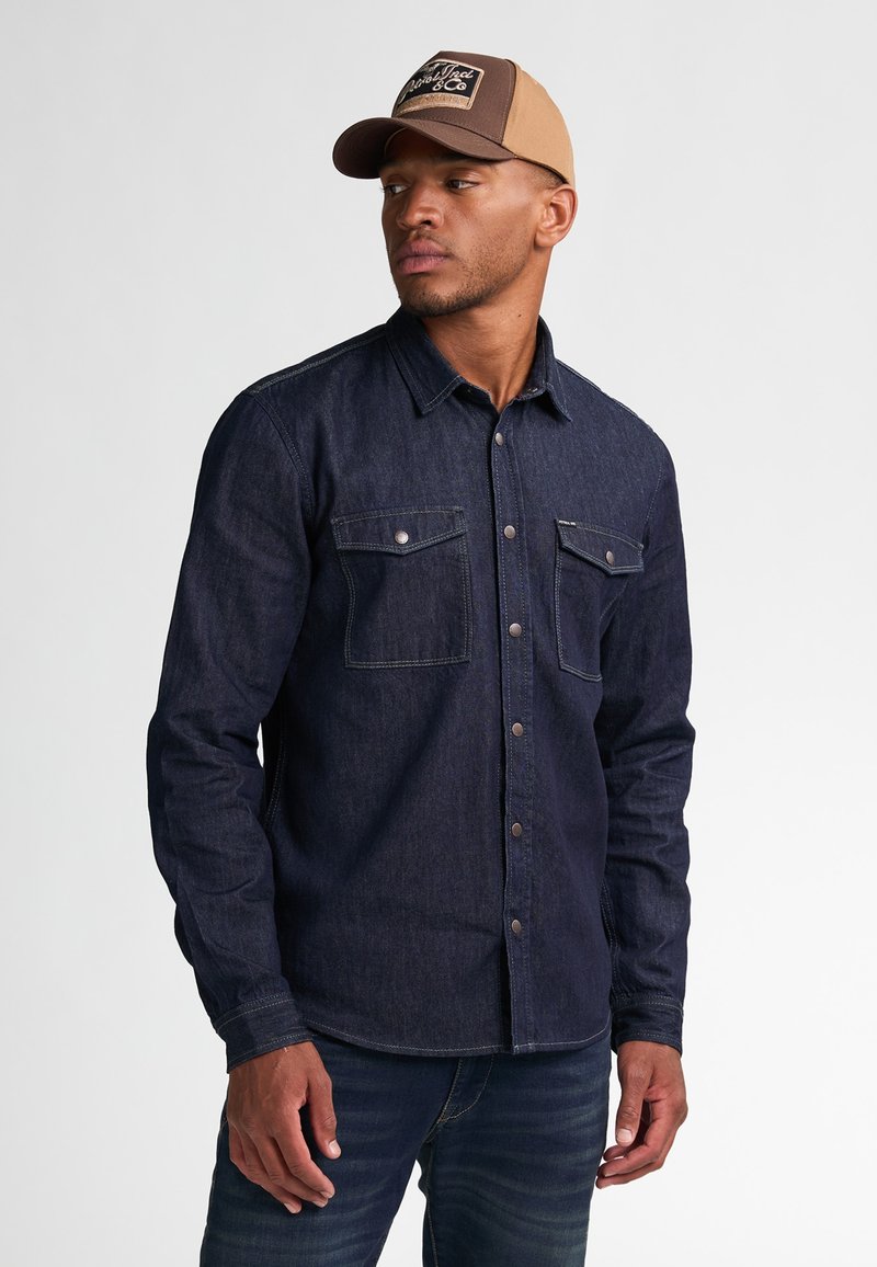 Camisa de mezclilla oscura de botones con mangas largas y dos bolsillos en el pecho, combinada con jeans oscuros. El modelo lleva una gorra marrón con un logo.