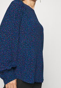 Blusa a fantasia blu e viola con maniche lunghe e gonfie. Tessuto morbido e vestibilità ampia, con scollatura rotonda e silhouette rilassata.