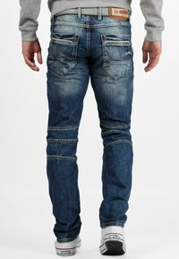 Jean tapered bleu foncé en denim, avec des zones décolorées, plusieurs poches arrière et une étiquette de marque en cuir à la taille.
