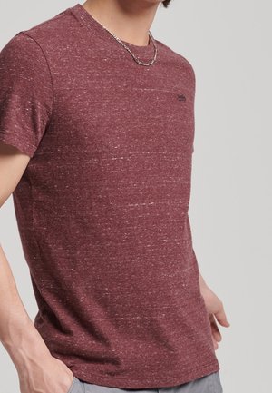 Homme portant un t-shirt à manches courtes bordeaux moucheté et un collier en chaîne argenté, debout devant un fond clair neutre.