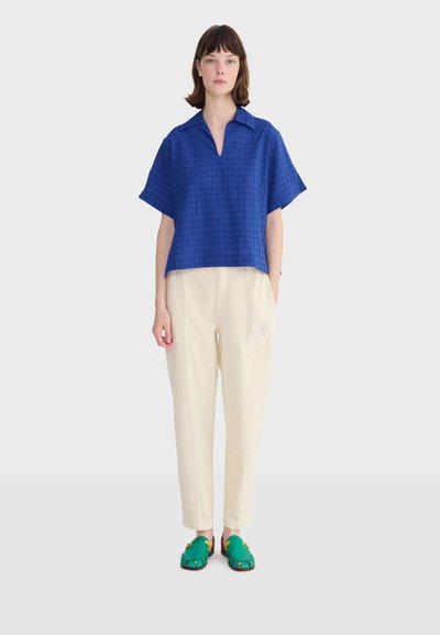 Femme debout portant un haut ample à manches courtes bleu, un pantalon crème fuselé et des sandales vertes à bouts ouverts, sur un fond clair uni.