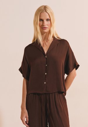 Donna bionda che indossa una camicia marrone scuro a maniche corte, larga e abbinata a pantaloni, in piedi davanti a uno sfondo semplice.