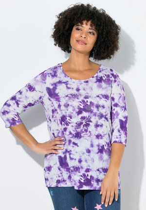 Kobieta z kręconymi włosami ubrana w fioletową koszulkę tie-dye z wzorem z dżetów i niebieskie jeansy z wzorem gwiazd, pozująca na tle białej ściany.