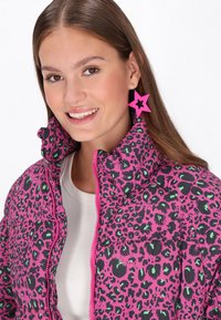 Jachetă pufosă roz cu imprimeu leopard negru și verde, guler înalt și închidere cu fermoar. Modelul poartă cercei mari în formă de stea roz.