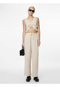 Beige kort topp med framtill rynkning och justerbara snören, parat med vida beige byxor. Modellen håller en liten svart väska och bär svarta solglasögon.