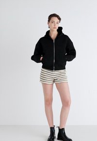 Svart zip-up hoodie med huva, bärs över krämfärgade shorts med svarta horisontella ränder. Modellen har på sig svarta ankelstövlar.