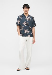 Homme portant une chemise à manches courtes bleu foncé à motif floral, un pantalon blanc à jambes larges et des tongs noires, debout les mains dans les poches sur fond blanc.