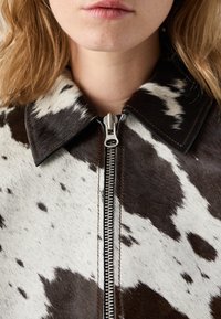 Braune und weiße Kuhfell-Jacke mit Reißverschluss, spitzem Kragen und metallischem Reißverschluss. Zeigt eine Mischung aus glatten und strukturierten Oberflächen.
