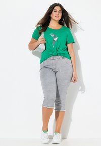 Camiseta de algodón verde con un diseño de nudo en la cintura, que presenta un gráfico colorido. Combinada con pantalones capri grises y zapatillas blancas.