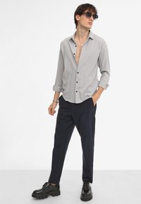 Camisa de botones de color gris claro con un patrón texturizado, mangas enrolladas, botones negros, combinada con pantalones oscuros y zapatos de plataforma negros y brillantes.
